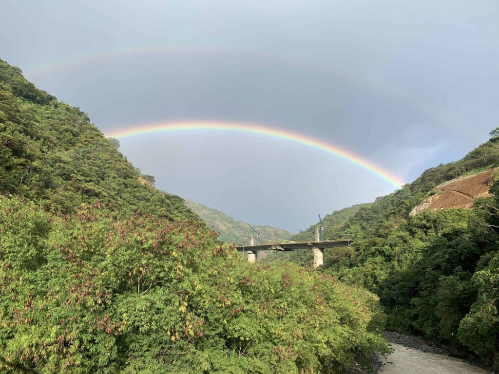 Un impresionante paisaje de un río que fluye entre majestuosas montañas, con un arcoíris vibrante que atraviesa el cielo. Trabajadores de la construcción están construyendo un puente, simbolizando el progreso y la unión entre la naturaleza colombiana y el empuje colombiano